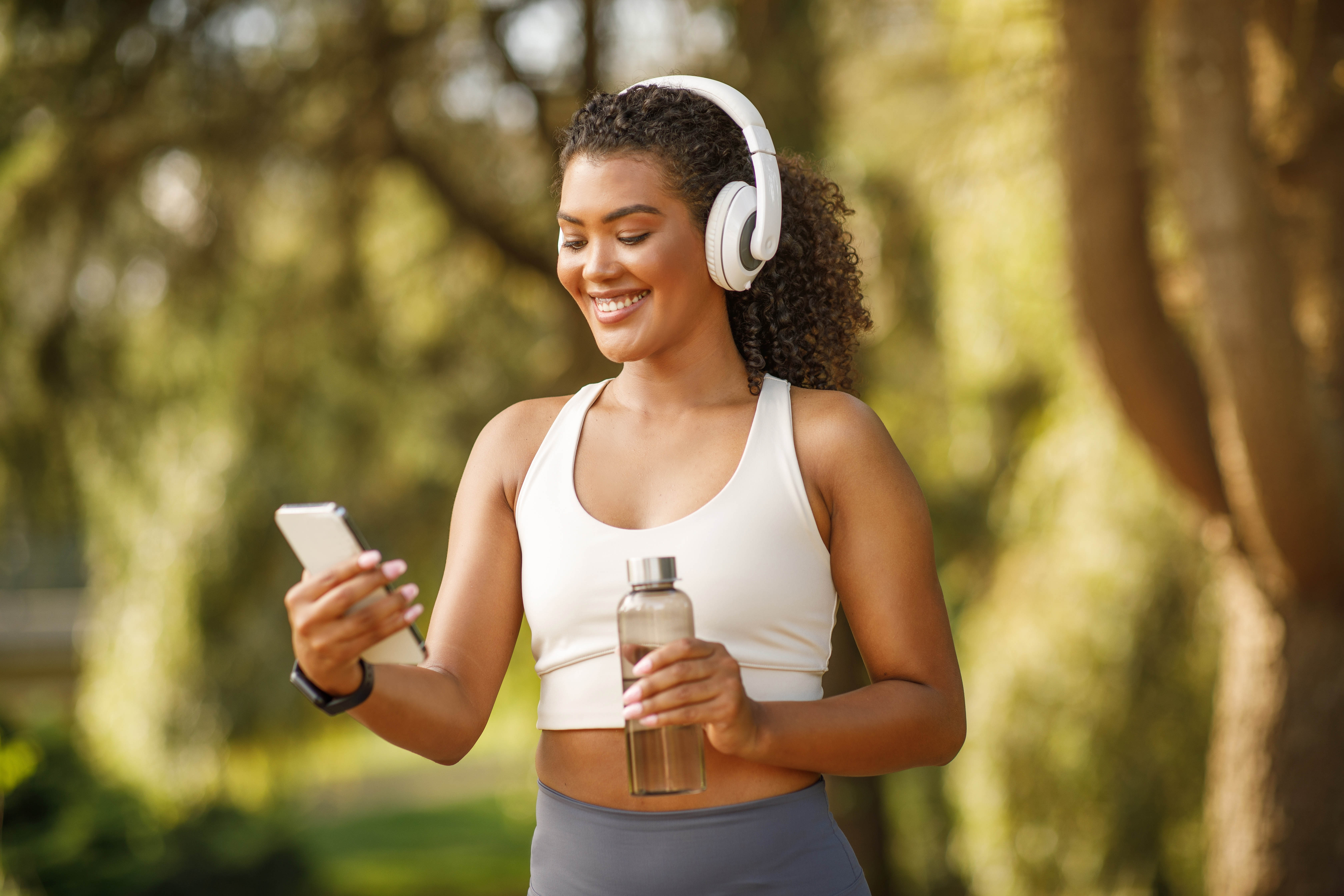 Young woman outside with her headphones on, smiling at her phone doing an outdoor workout